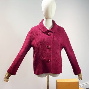 May include: A magenta-colored jacket with a round collar and three matching buttons. The jacket is displayed on a mannequin with a gold stand. The jacket has a textured fabric and a classic design.
