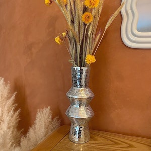 May include: A silver mirrored vase with a geometric design holds a bouquet of dried yellow flowers and wheat stalks.