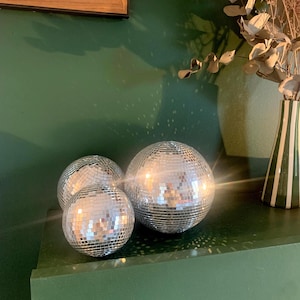 Op de afbeelding: Drie bolvormige discoballen bedekt met kleine, reflecterende zilveren spiegeltjes. De bollen variëren in grootte en zijn in een groepje gerangschikt, waarbij ze licht weerkaatsen en heldere schitteringen creëren op hun gefacetteerde oppervlakken.