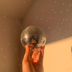 Puede incluir: Una mano sosteniendo una pequeña bola de discoteca. La bola está cubierta de pequeñas baldosas de espejo cuadradas que reflejan la luz. El fondo es de un color cálido y neutro con pequeños puntos blancos, creando un efecto de estrellas. La iluminación sugiere un ambiente cálido y soleado.
