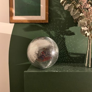 Op de afbeelding: Een zilveren discobal ligt op een donkergroen oppervlak en reflecteert licht. De bal is rond en bedekt met kleine spiegelvierkantjes. Een ingelijst kunstwerk en een vaas met gedroogde bloemen zijn op de achtergrond.