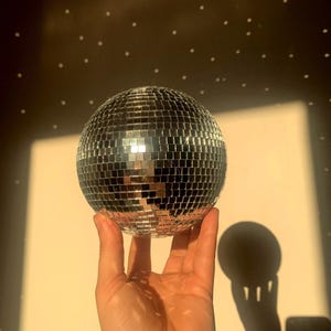 Könnte beinhalten: Eine silberne Discokugel mit verspiegelter Oberfläche wird von einer Hand gehalten. Der Ball reflektiert Licht und erzeugt einen funkelnden Effekt. Der Hintergrund ist eine warme, neutrale Farbe mit einem Muster aus kleinen weißen Punkten.