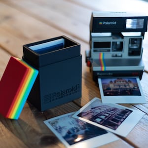 May include: A vintage Polaroid camera with a rainbow stripe, next to a black box of film and a stack of colorful film packs. Two printed photos are visible. The box has the Polaroid logo.