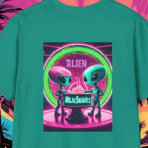May include: A teal green t-shirt with a graphic print of two green aliens holding milkshakes. The graphic is set against a neon pink and blue background with the text "Alien Milkshakes".