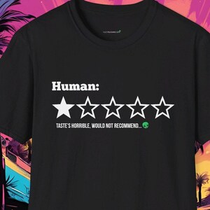 May include: Black t-shirt with a white graphic of a one-star review for "Human." The text reads "Taste's horrible, would not recommend..." with a green alien emoji.