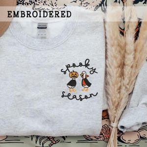 K&ouml;nnte beinhalten: Hellgraues Sweatshirt mit gesticktem Schriftzug "spooky season" und zwei Cartoon-Figuren: einem K&uuml;rbis und einer Vampir-Ente. Das Wort "EMBROIDERED" steht &uuml;ber dem Sweatshirt.