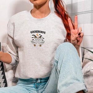 May include: Light gray sweatshirt with the word "GOOSEBUMPS" above an embroidered design of two geese wearing witch hats. The sweatshirt is paired with light blue jeans.