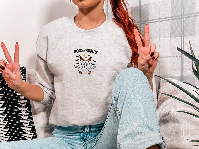 May include: Light gray sweatshirt with the word "GOOSEBUMPS" above an embroidered design of two geese wearing witch hats. The sweatshirt is paired with light blue jeans.