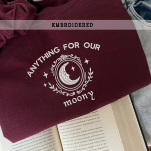 Könnte beinhalten: Burgunderfarbenes Sweatshirt mit weiß gesticktem Text und einem Mond-Design. Der Text lautet "ANYTHING FOR OUR moony" und "EMBROIDERED". Das Design zeigt einen Mond, Sterne und dekorative Verzierungen.