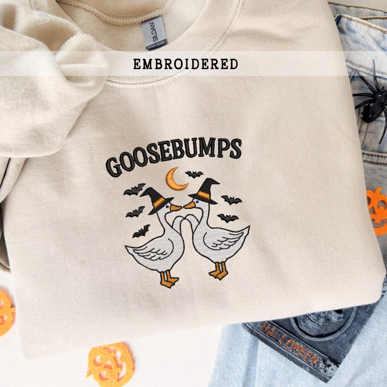 May include: Cream-colored embroidered sweatshirt with the word "GOOSEBUMPS" in black. The design features two geese wearing witch hats, a crescent moon, and bats. The word "EMBROIDERED" is above the main design.