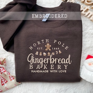 Może przedstawiać: Brązowa haftowana bluza z napisem "North Pole Gingerbread Bakery" w kremowym kolorze. Projekt zawiera piernikowych ludzików i inne świąteczne kształty. Bluza jest złożona na białej powierzchni z dekoracjami świątecznymi.