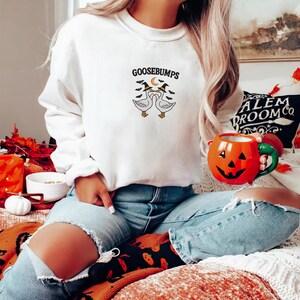 May include: White sweatshirt with the word "GOOSEBUMPS" above a graphic of two geese wearing witch hats. The geese are facing each other with a crescent moon between them. The person is wearing blue jeans and holding a pumpkin mug.