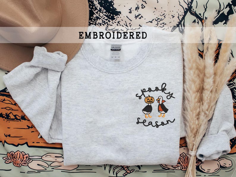 K&ouml;nnte beinhalten: Hellgraues Sweatshirt mit gesticktem Schriftzug "spooky season" und zwei Cartoon-Figuren: einem K&uuml;rbis und einer Vampir-Ente. Das Wort "EMBROIDERED" steht &uuml;ber dem Sweatshirt.