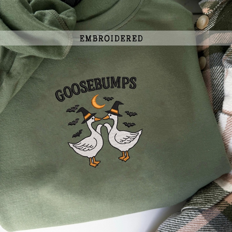 May include: An olive green sweatshirt with the word "GOOSEBUMPS" embroidered in black. The design features two white geese wearing witch hats, a crescent moon, and bats. The word "EMBROIDERED" is above the design.