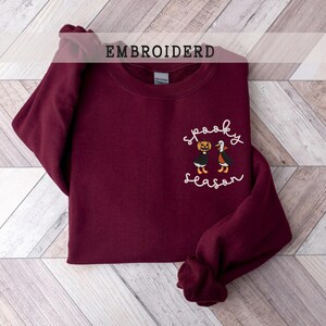 K&ouml;nnte beinhalten: Ein weinrotes Sweatshirt mit dem gestickten Schriftzug "Spooky Season" und einer Grafik einer K&uuml;rbis und einer Fledermaus. Das Wort "EMBROIDERD" ist oben im Bild aufgedruckt. Das Sweatshirt liegt gefaltet auf einer hellen Holzmaserung.