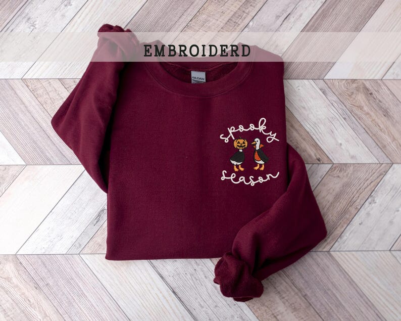 K&ouml;nnte beinhalten: Ein weinrotes Sweatshirt mit dem gestickten Schriftzug "Spooky Season" und einer Grafik einer K&uuml;rbis und einer Fledermaus. Das Wort "EMBROIDERD" ist oben im Bild aufgedruckt. Das Sweatshirt liegt gefaltet auf einer hellen Holzmaserung.