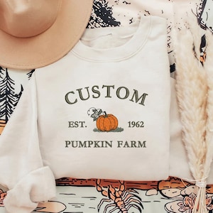 Peut inclure: Sweat-shirt crème avec l'inscription "CUSTOM PUMPKIN FARM EST. 1962" en vert. Le motif comprend une illustration de citrouille orange. Un chapeau beige est visible en haut à gauche.