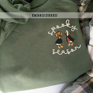 K&ouml;nnte beinhalten: Olivgr&uuml;nes, besticktes Sweatshirt mit den Worten "spooky season" und einer K&uuml;rbis- und Pinguin-Grafik. Das Wort "EMBROIDERED" steht &uuml;ber der Grafik. Das Sweatshirt ist gefaltet.