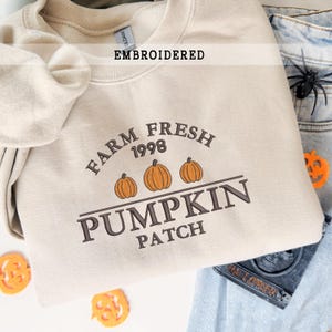 Könnte beinhalten: Beiges besticktes Sweatshirt mit dem Text "Farm Fresh Pumpkin Patch 1998" und drei orangefarbenen Kürbissen. Das Wort "EMBROIDERED" steht über dem Design. Das Sweatshirt ist gefaltet, mit einer Jeans und Halloween-Dekorationen im Hintergrund.
