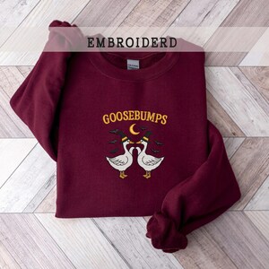 May include: A maroon sweatshirt with the word "GOOSEBUMPS" embroidered in yellow, with a crescent moon and two geese wearing hats. The word "EMBROIDERD" is printed at the top. The sweatshirt is folded on a light wood-grain surface.