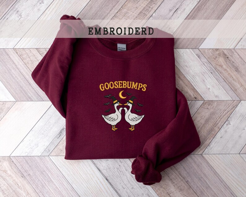 May include: A maroon sweatshirt with the word "GOOSEBUMPS" embroidered in yellow, with a crescent moon and two geese wearing hats. The word "EMBROIDERD" is printed at the top. The sweatshirt is folded on a light wood-grain surface.