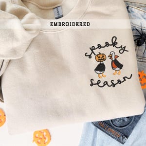 K&ouml;nnte beinhalten: Beige besticktes Sweatshirt mit dem Schriftzug "Spooky Season" und zwei Halloween-Charakteren. Einer ist ein K&uuml;rbis, der andere eine Ente mit Umhang. Das Wort "EMBROIDERED" steht &uuml;ber dem Kragen.