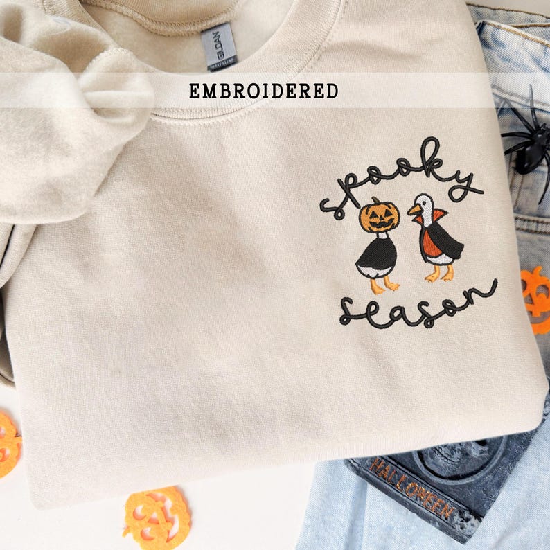 K&ouml;nnte beinhalten: Beige besticktes Sweatshirt mit dem Schriftzug "Spooky Season" und zwei Halloween-Charakteren. Einer ist ein K&uuml;rbis, der andere eine Ente mit Umhang. Das Wort "EMBROIDERED" steht &uuml;ber dem Kragen.