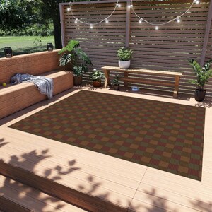 Op de afbeelding: Een geruit buitenkleed met bruine, groene en rode vierkanten. Het kleed ligt op een houten dek met een houten bank en planten op de achtergrond.