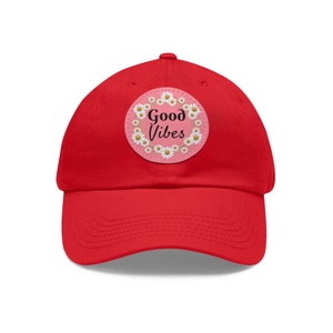 Peut inclure: Casquette de baseball rouge avec un patch de marguerite rose et blanche qui dit "Good Vibes".