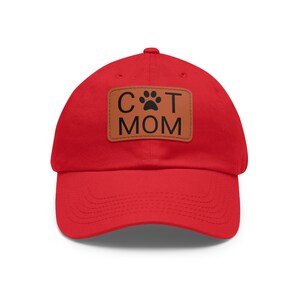 Peut inclure: Casquette de baseball rouge avec un patch en cuir marron qui dit "CAT MOM" avec une empreinte de patte.