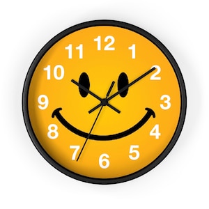 Pode incluir: Um relógio de parede redondo com moldura preta e mostrador amarelo com um design de carinha feliz. O relógio tem ponteiros pretos e algarismos brancos. A carinha feliz tem olhos pretos e uma boca preta curvada.