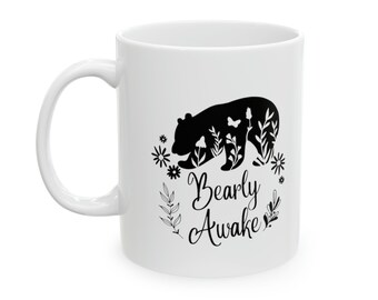 Op de afbeelding: Witte keramische mok met een zwart silhouet van een beer omringd door bloemen en bladeren. De tekst "Bearly Awake" staat onder de beer.