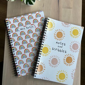 May include: Two spiral-bound notebooks with white covers. One features a pattern of colorful rainbows and the text "Wee Notes." The other has a design of yellow and peach suns with the words "notes and scribbles."