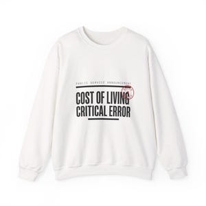 May include: White sweatshirt featuring the text "PUBLIC SERVICE ANNOUNCEMENT" above "COST OF LIVING CRITICAL ERROR" in black, with a red stamp graphic. The sweatshirt has a crew neck and long sleeves.