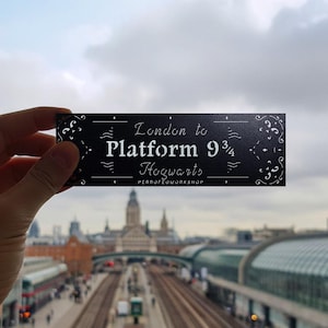 Puede incluir: Un letrero rectangular negro con texto blanco que dice "London to Platform 9 3/4 Hogwarts". El letrero tiene adornos decorativos y se muestra frente a un paisaje urbano borroso con vías de tren.