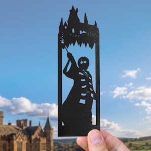 Puede incluir: Un marcapáginas de metal negro con la silueta cortada a láser de un mago con gafas redondas y bufanda a rayas, sosteniendo una varita en alto. Sobre la figura hay un recorte detallado de un castillo con varias torres y agujas.
