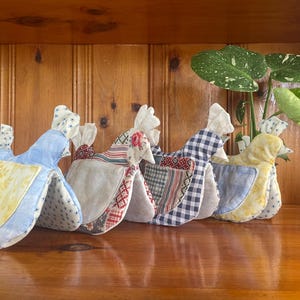 May include: A row of four fabric chicken-shaped kitchen accessories. Each piece features a unique combination of patterned textiles, including blue, yellow, red, and white gingham, floral, and geometric prints, with a distinct pocket on the side.