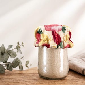 Peut inclure: Un bocal en verre transparent rempli d'une substance brun clair, surmonté d'un couvercle en tissu. Le couvercle présente un motif de fraises rouges, de citrons jaunes et de feuilles vertes sur fond crème. Le bocal est posé sur une surface en bois.