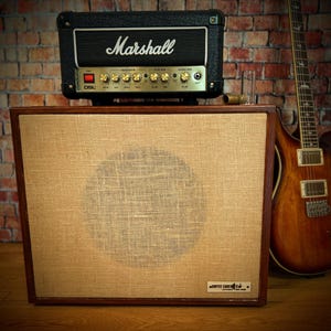 May include: A black Marshall amplifier sits atop a large speaker cabinet with a tan fabric front. A brown electric guitar leans against a brick wall. The speaker cabinet has a dark wood frame and a circular design on the front.