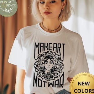 Puede incluir: Camiseta blanca con un estampado gráfico negro que dice "Make Art Not War". El gráfico presenta el rostro de una mujer con flores a su alrededor. La camiseta está siendo modelada por una persona.
