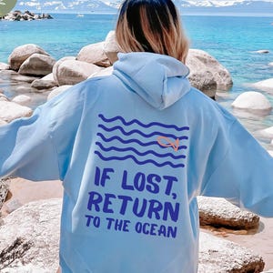 May include: Light blue hoodie with the text "IF LOST, RETURN TO THE OCEAN" and a graphic of waves and a swimmer. The hoodie has a drawstring hood and long sleeves. The background shows a beach with rocks and water.