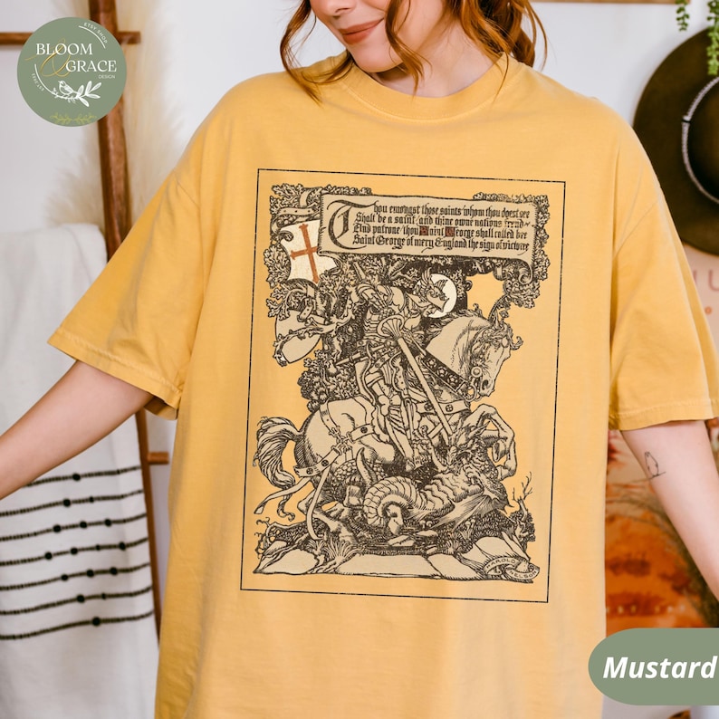 Pu&ograve; includere: T-shirt color senape con un'illustrazione dettagliata in bianco e nero di un cavaliere a cavallo che uccide un drago, con testo sopra. La maglietta ha un girocollo e maniche corte.
