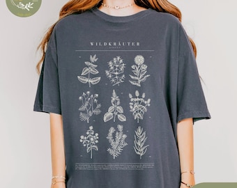 Puede incluir: Camiseta de color denim con una ilustración botánica de varias flores silvestres. La camiseta tiene mangas cortas y un corte holgado. El texto "WILDKRAUTER" está impreso en la parte superior del diseño. La palabra "Denim" está impresa en la esquina inferior derecha.
