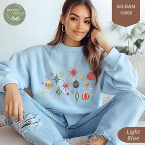 Puede incluir: Una sudadera de color azul claro con un diseño de adornos navideños. El diseño presenta adornos rojos, verdes y dorados con estrellas. La sudadera está hecha por Gildan y tiene el número de estilo 18000.