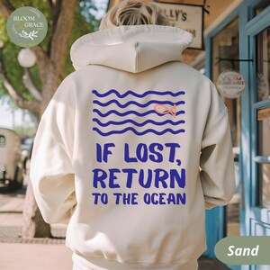 May include: Sand-colored hoodie with a blue and orange graphic on the back. The graphic features blue wavy lines, an orange fish, and the text "IF LOST, RETURN TO THE OCEAN" in blue. The hoodie has a hood and a drawstring.