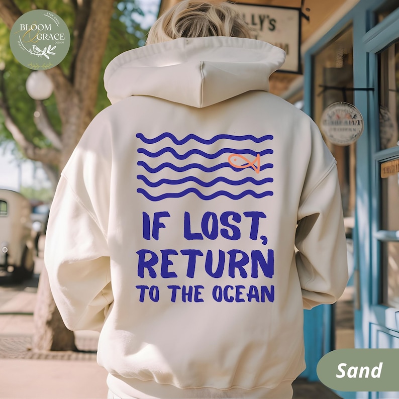 May include: Sand-colored hoodie with a blue and orange graphic on the back. The graphic features blue wavy lines, an orange fish, and the text "IF LOST, RETURN TO THE OCEAN" in blue. The hoodie has a hood and a drawstring.