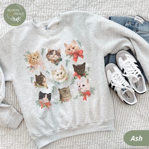 Peut inclure: Sweat-shirt gris cendr&eacute; avec un motif de visages de chats orn&eacute;s de n&oelig;uds rouges et de verdure. Le sweat-shirt est pr&eacute;sent&eacute; avec un jean bleu et des baskets blanches &agrave; rayures noires. Le logo Bloom & Grace est visible.