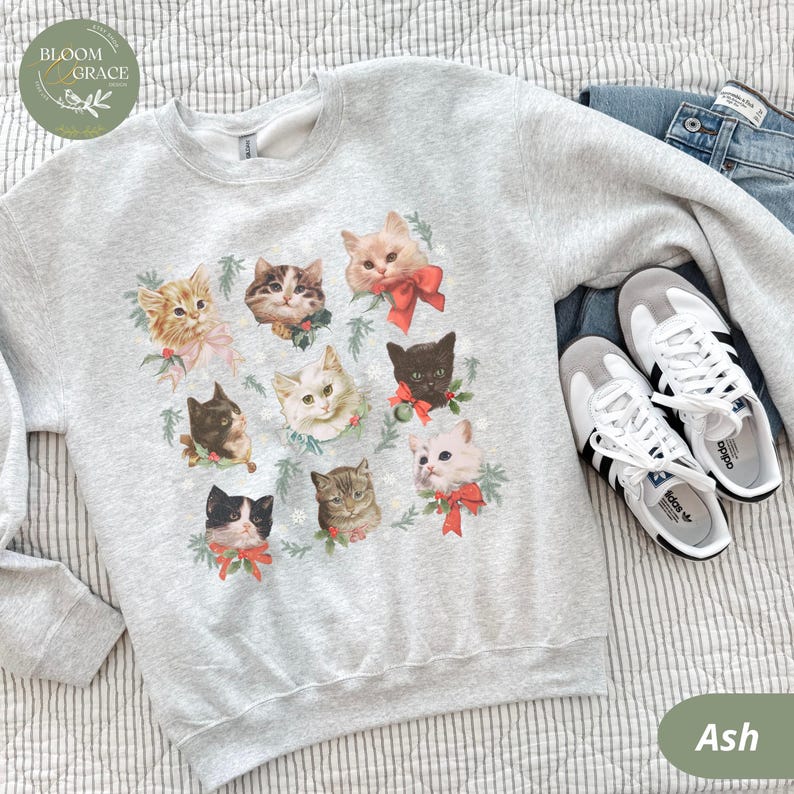 Peut inclure: Sweat-shirt gris cendr&eacute; avec un motif de visages de chats orn&eacute;s de n&oelig;uds rouges et de verdure. Le sweat-shirt est pr&eacute;sent&eacute; avec un jean bleu et des baskets blanches &agrave; rayures noires. Le logo Bloom & Grace est visible.