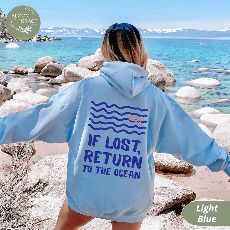 May include: Light blue hoodie with the text "IF LOST, RETURN TO THE OCEAN" and a graphic of waves and a swimmer. The hoodie has a drawstring hood and long sleeves. The background shows a beach with rocks and water.