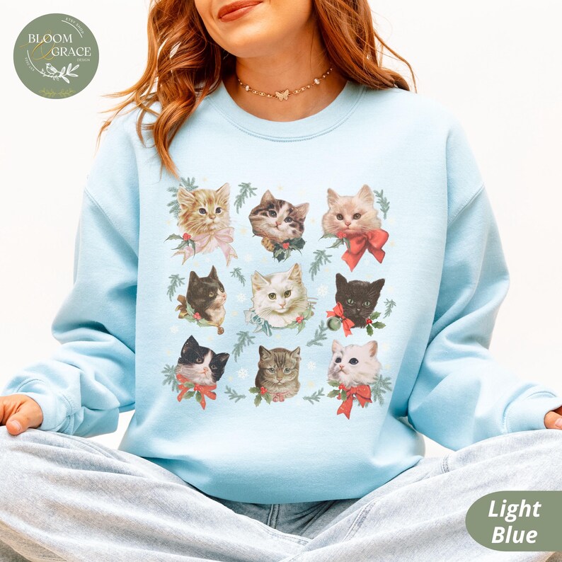 Peut inclure: Sweat-shirt bleu clair orn&eacute; d'une grille de neuf portraits de chats, chacun orn&eacute; d'un n&oelig;ud et de verdure. Les chats varient en couleur et en motif, sur fond bleu clair. Le texte "Bloom & Grace" est dans un cercle en haut &agrave; gauche.